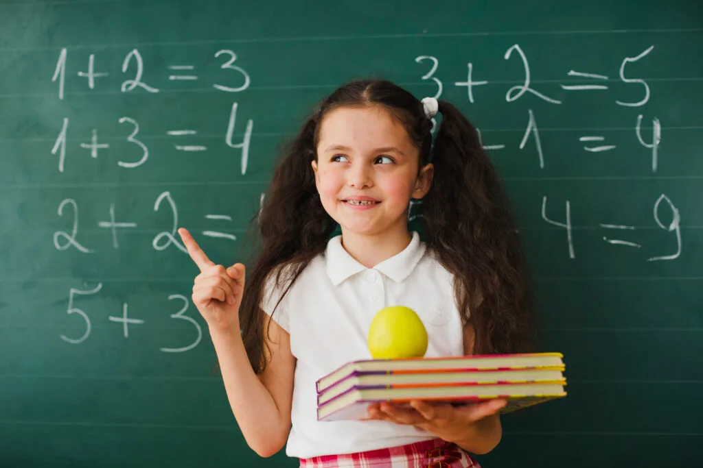 Smiling schoolgirl holding books and pointing at math equations on the blackboard, symbolizing Vedic maths learning.